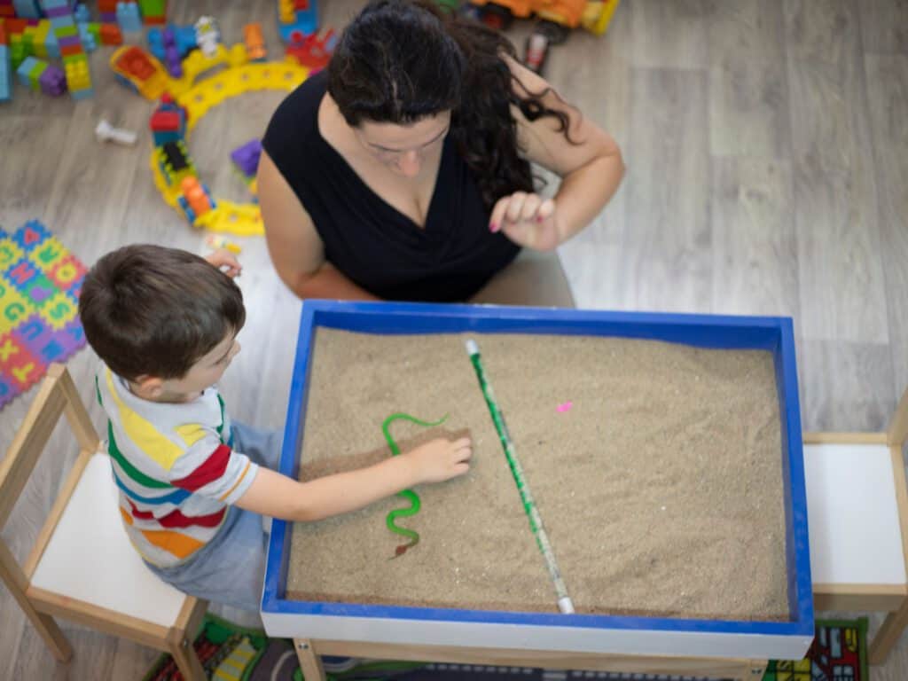 מטפלת וילד מעל שולחן חול קינטי, הילד מכניס את ידו בחול. ב INDEKIDS תוכלו למצוא מגוון של מטפלים במשחק