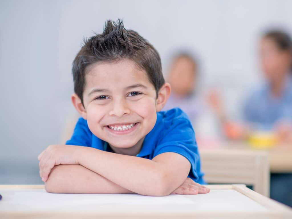 ילד בכיתה א מחייך יושב על שולחן בית הספר. ב-INDEKIDS תקבלו כלים על התמודדות עם המעבר מגן לכיתה א